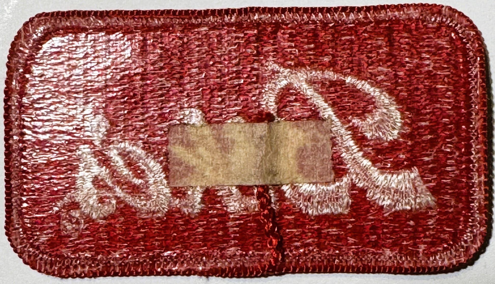 Rectangular red embroidered patch with white decorative scalloped border design and tan fabric label in center, photographed 