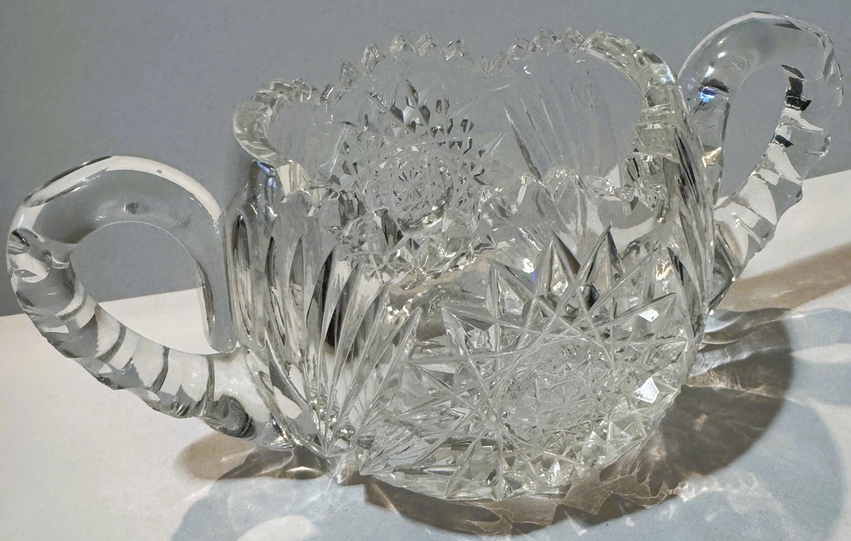 Antique cut glass two-handled sugar bowl with intricate geometric patterns, photographed from above at an angle against a lig
