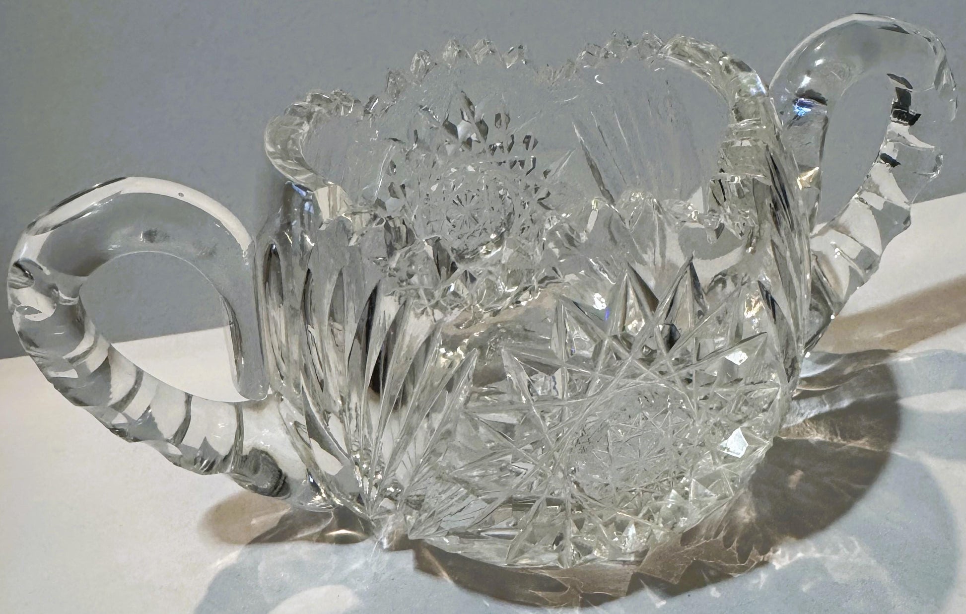 Antique cut glass two-handled sugar bowl with intricate geometric patterns, photographed from above at an angle against a lig