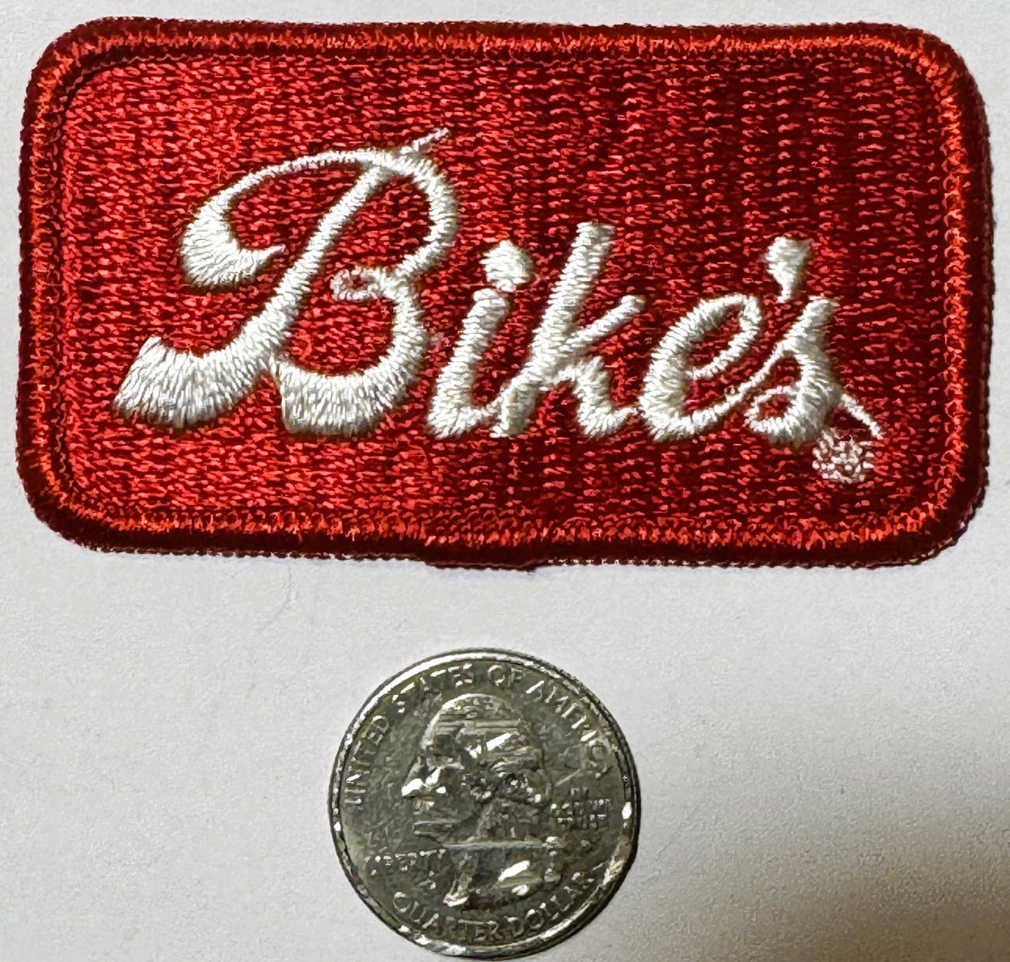 Red embroidered patch with white script "Bike's" lettering beside a quarter coin for scale