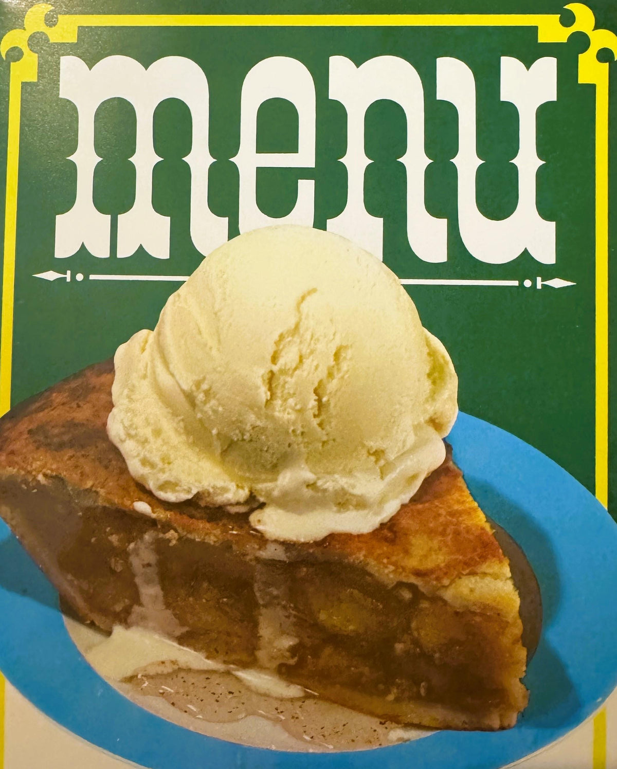 Close-up of the menu cover: big white menu letters on green above a scoop of vanilla ice cream melting on a slice of apple pie on a turquoise-rimmed plate