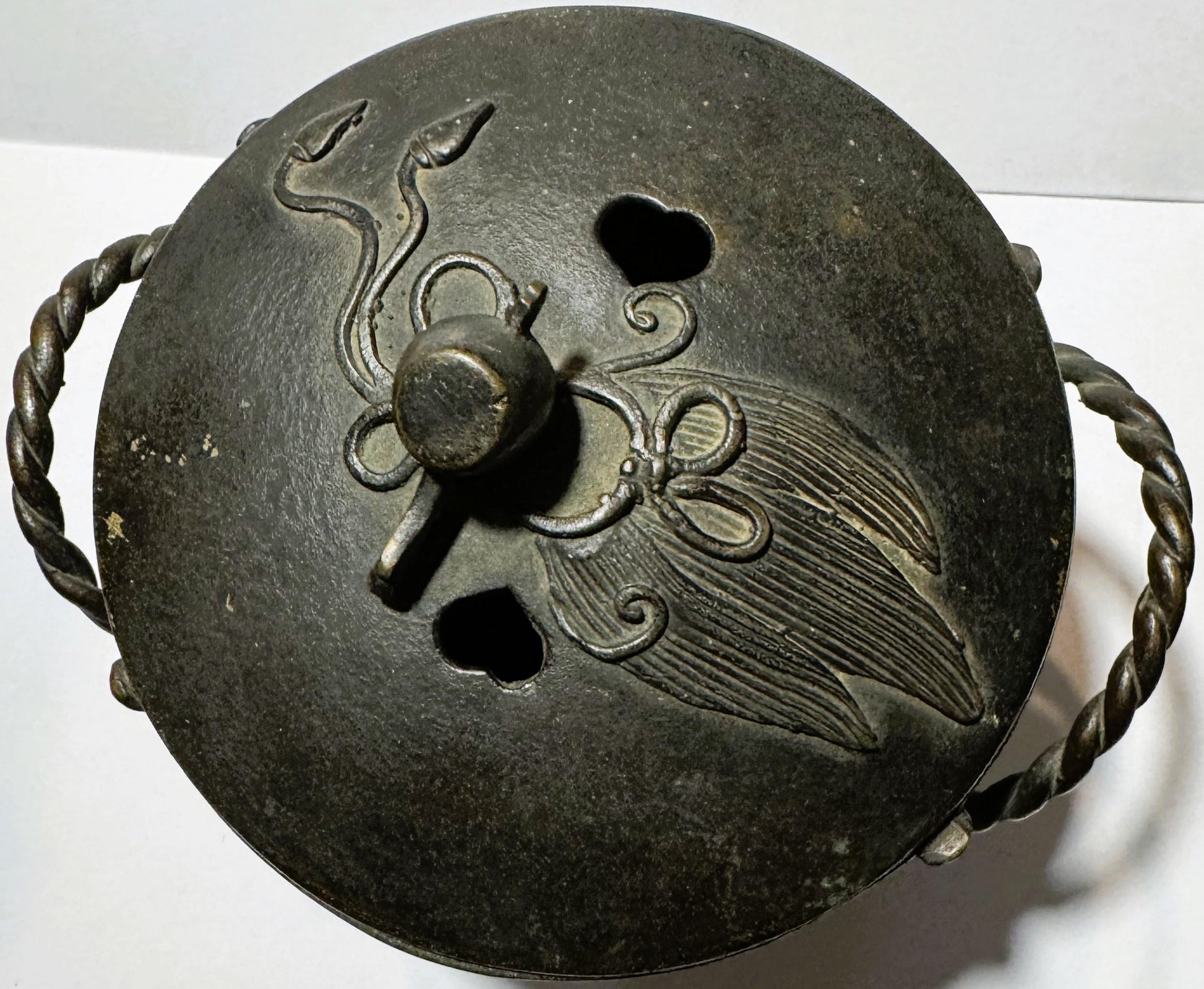 Top view of bronze censer lid with twisted handles, decorative scrollwork, central knob, and embossed bird design with detail