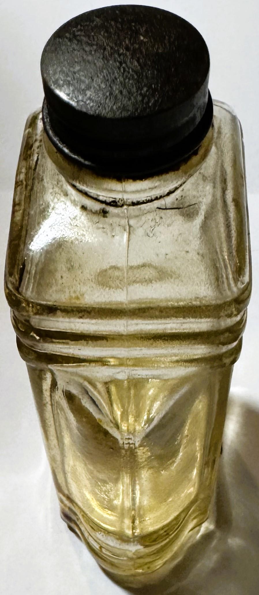 Vintage pressed glass bottle with yellowish liquid and black metal cap, viewed from front against white background, showing e