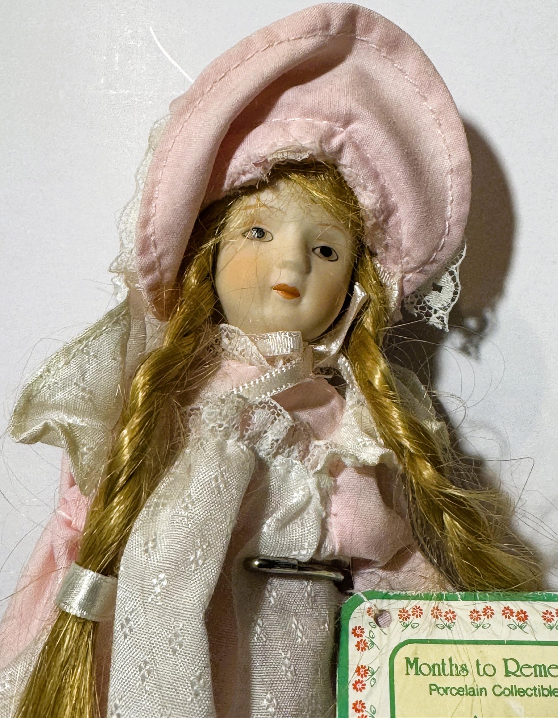 Porcelain doll with long blonde hair wearing pink bonnet and cream lace dress, with "Months to Remember" collectible tag visi