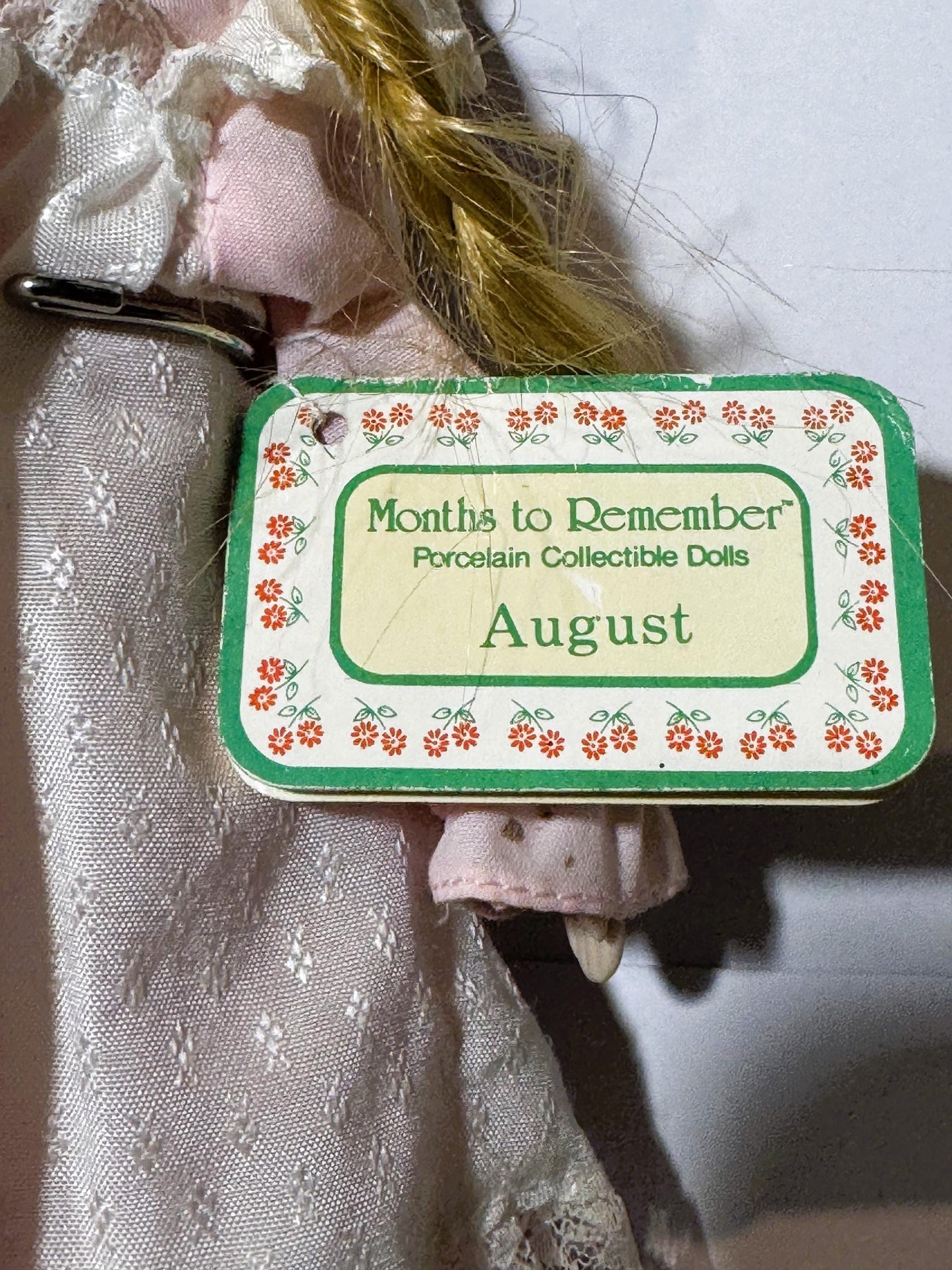 Close-up of green and cream labeled box for "Months to Remember Porcelain Collectible Dolls August" held against pink doll cl