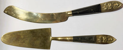 Two brass serving utensils with black handles and embossed Thai deity finials, displayed flat against white background