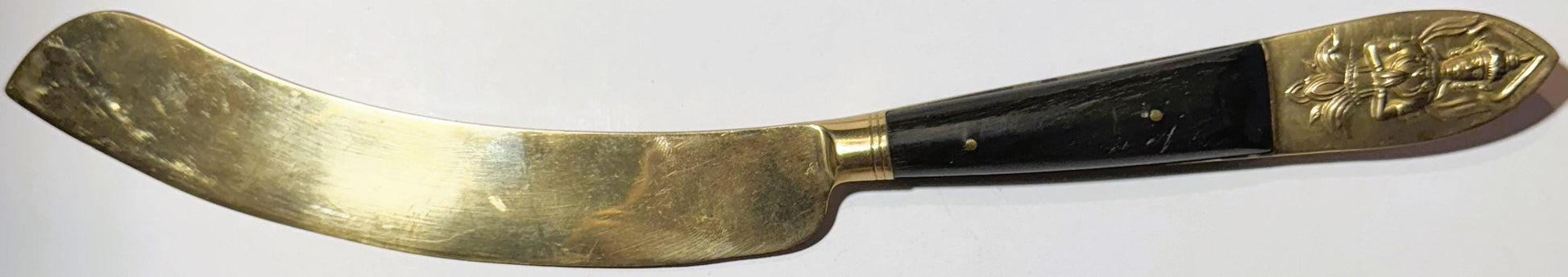 Antique brass serving knife with black handle and ornate Thai deity embossed on pommel, shown at angle against white backgrou