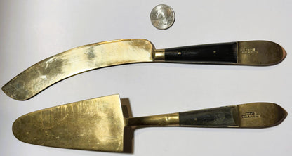 Two brass serving utensils with black handles displayed flat next to a quarter coin for scale, showing maker's marks on handl