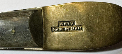 Close-up of brass belt buckle embossed "WILLY MADE IN SIAM" with dark wood backing, showing worn patina and surface texture