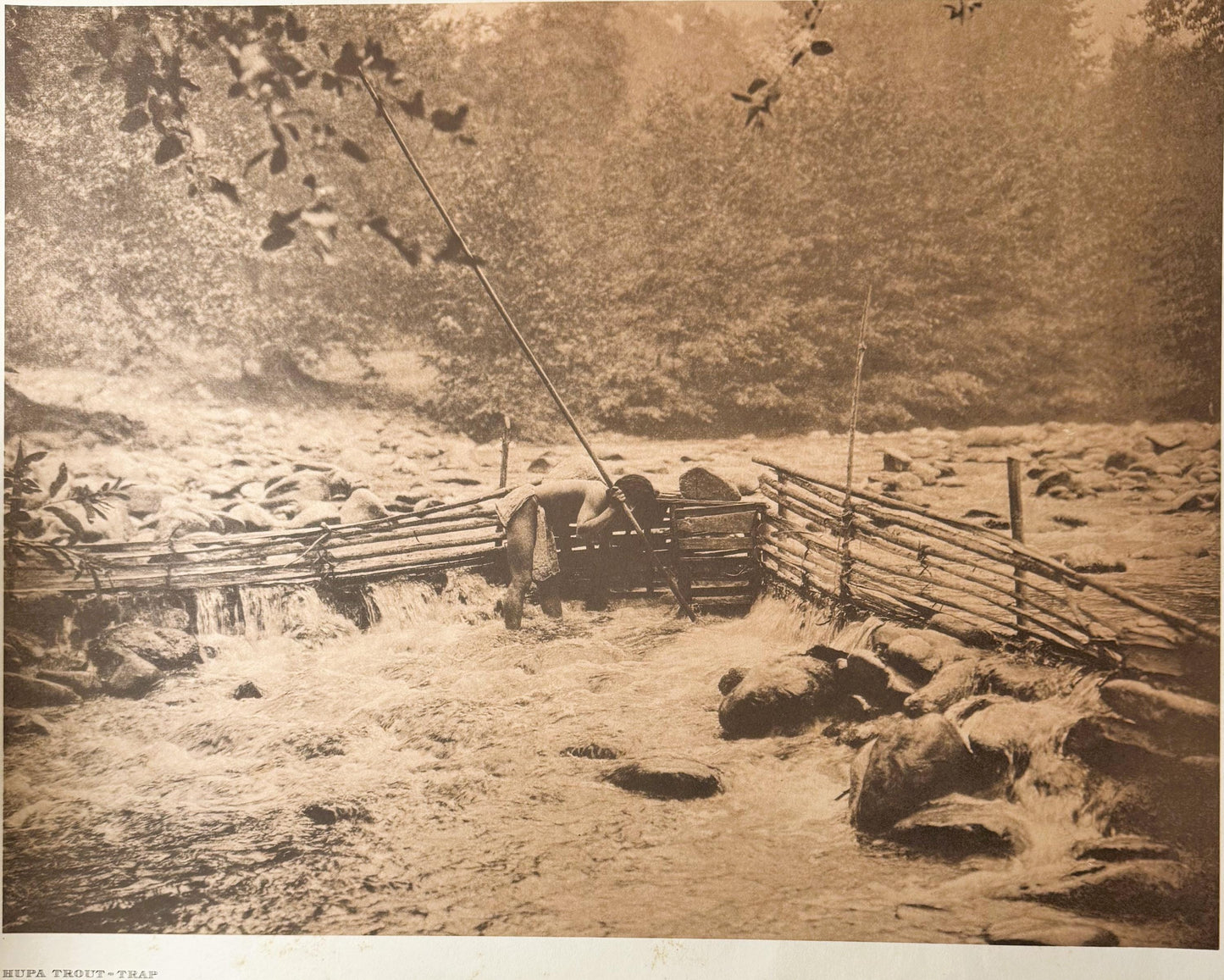 Sepia-toned photogravure showing a wooden fish trap structure spanning a rocky stream, with wooden fencing and forest backdro