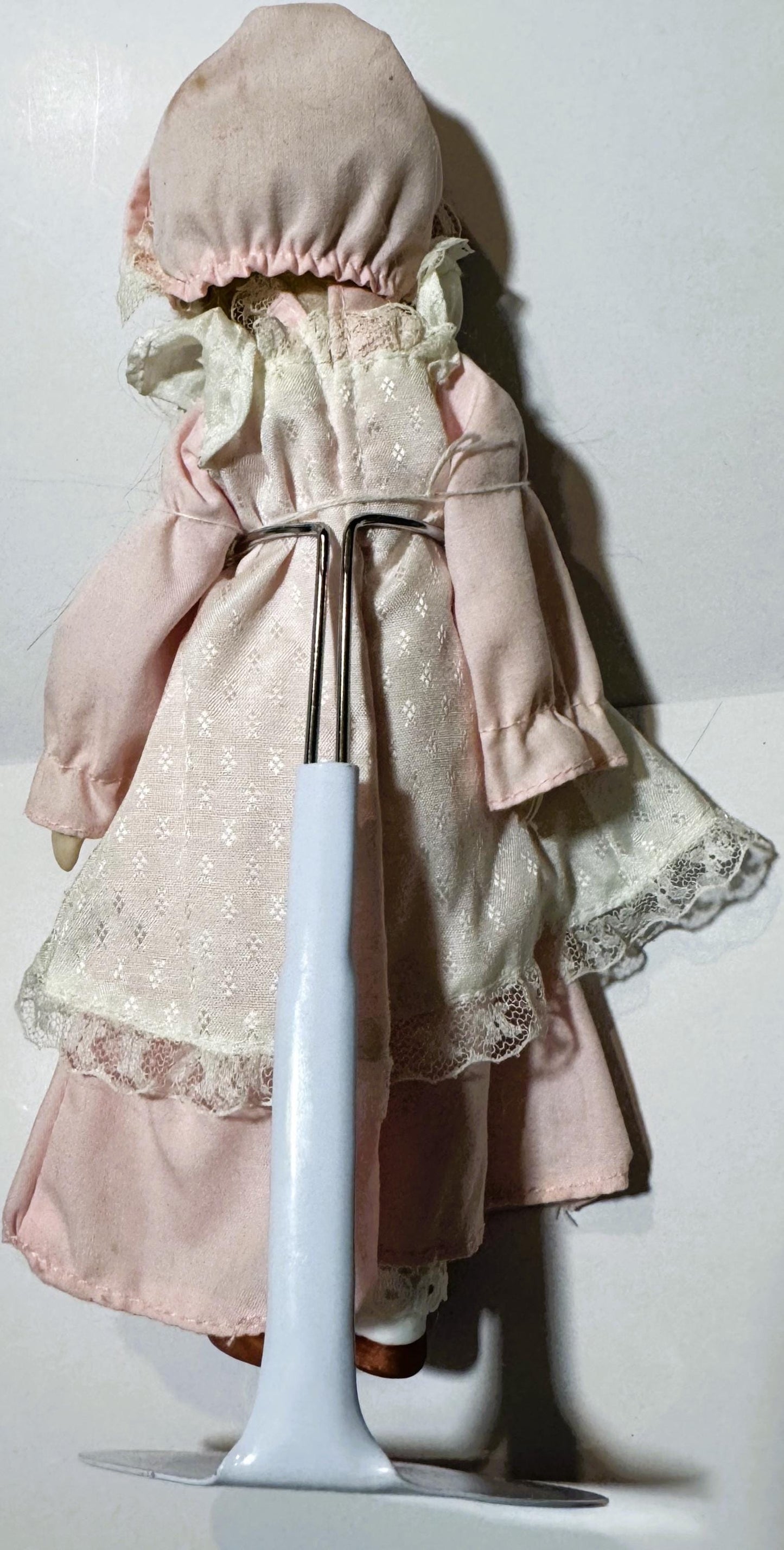 Vintage porcelain doll wearing pink dress and bonnet, shown from behind with white stand for scale