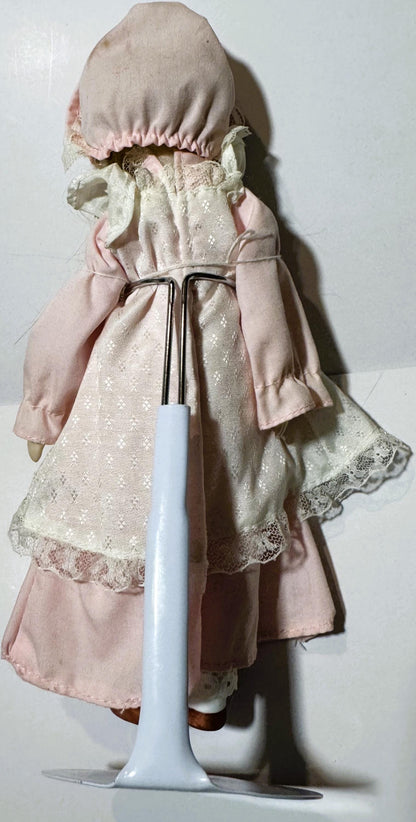 Vintage porcelain doll wearing pink dress and bonnet, shown from behind with white stand for scale