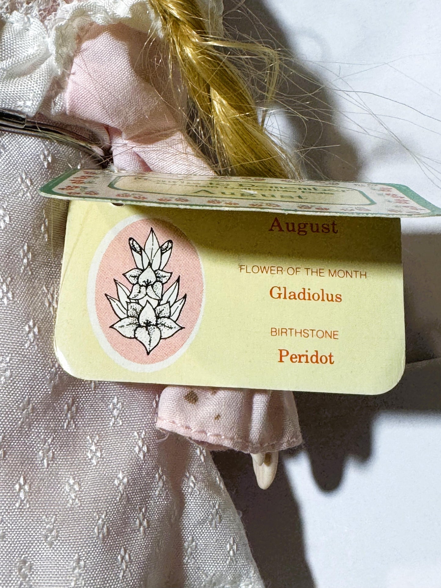 Close-up of vintage doll's tag reading "August / FLOWER OF THE MONTH / Gladiolus / BIRTHSTONE / Peridot" with gladiolus illus