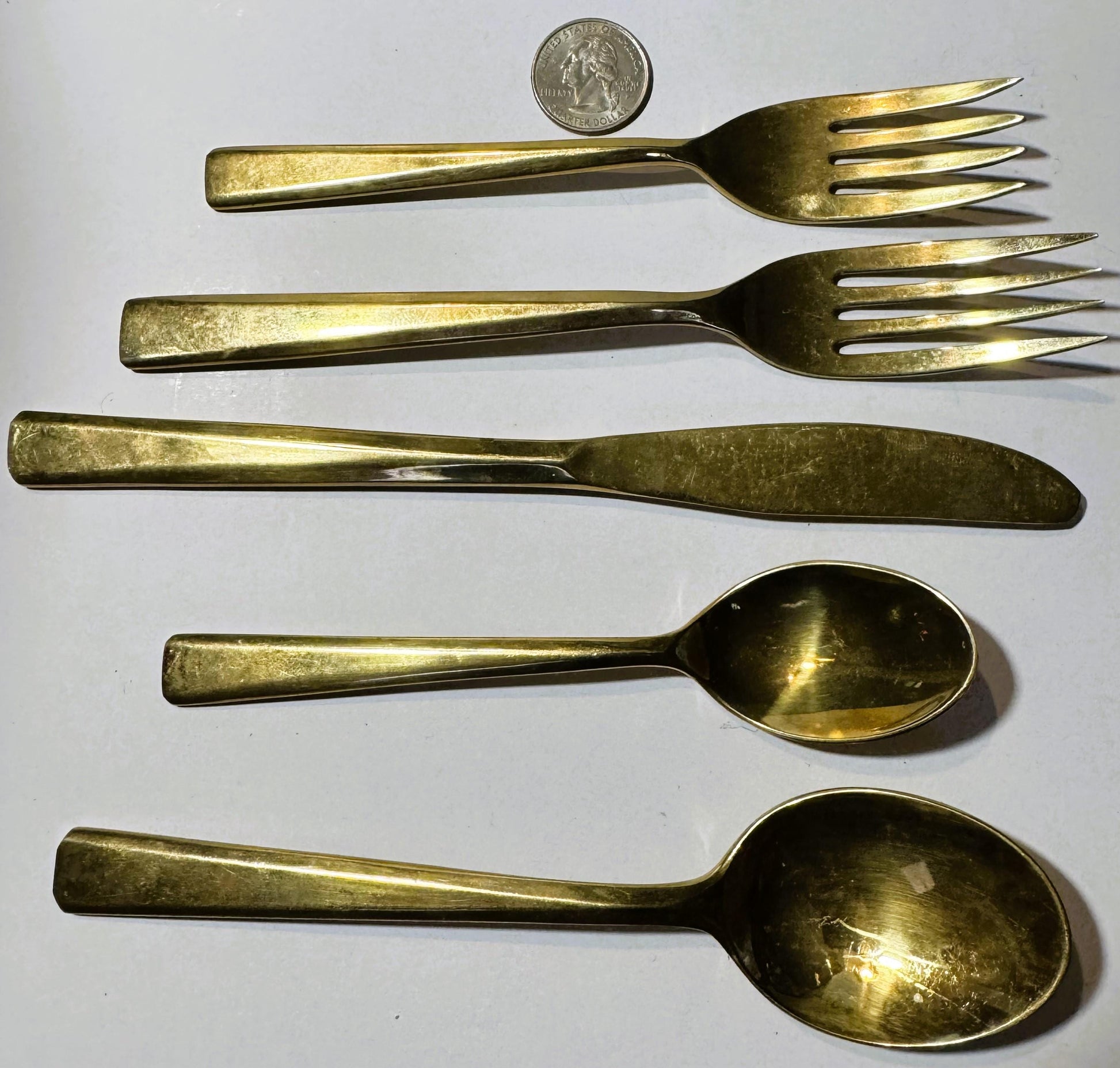 Five brass flatware pieces arranged vertically with a quarter coin at top for scale, showing two forks, knife, and two spoons