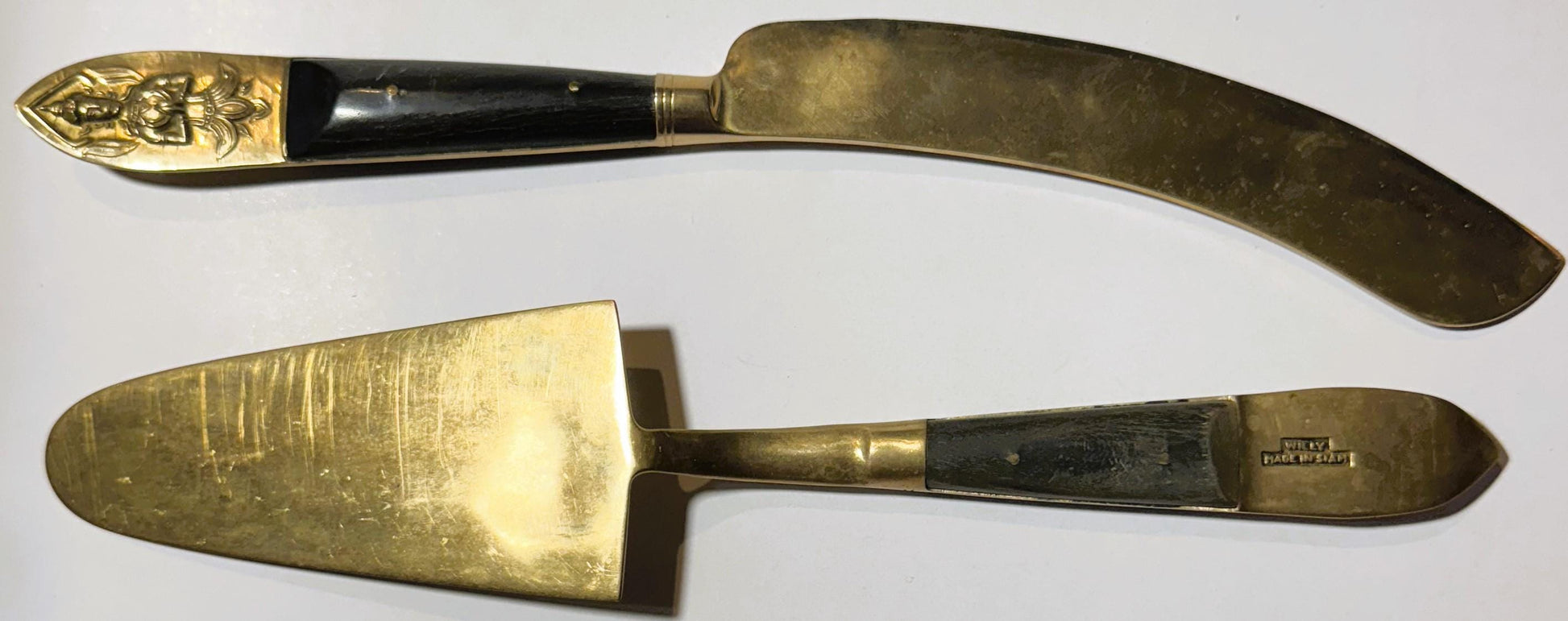 Two brass serving utensils with black handles displayed on white background: curved knife with embossed deity head handle and