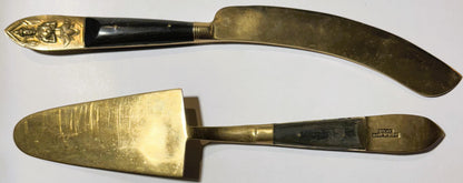 Two brass serving utensils with black handles displayed on white background: curved knife with embossed deity head handle and