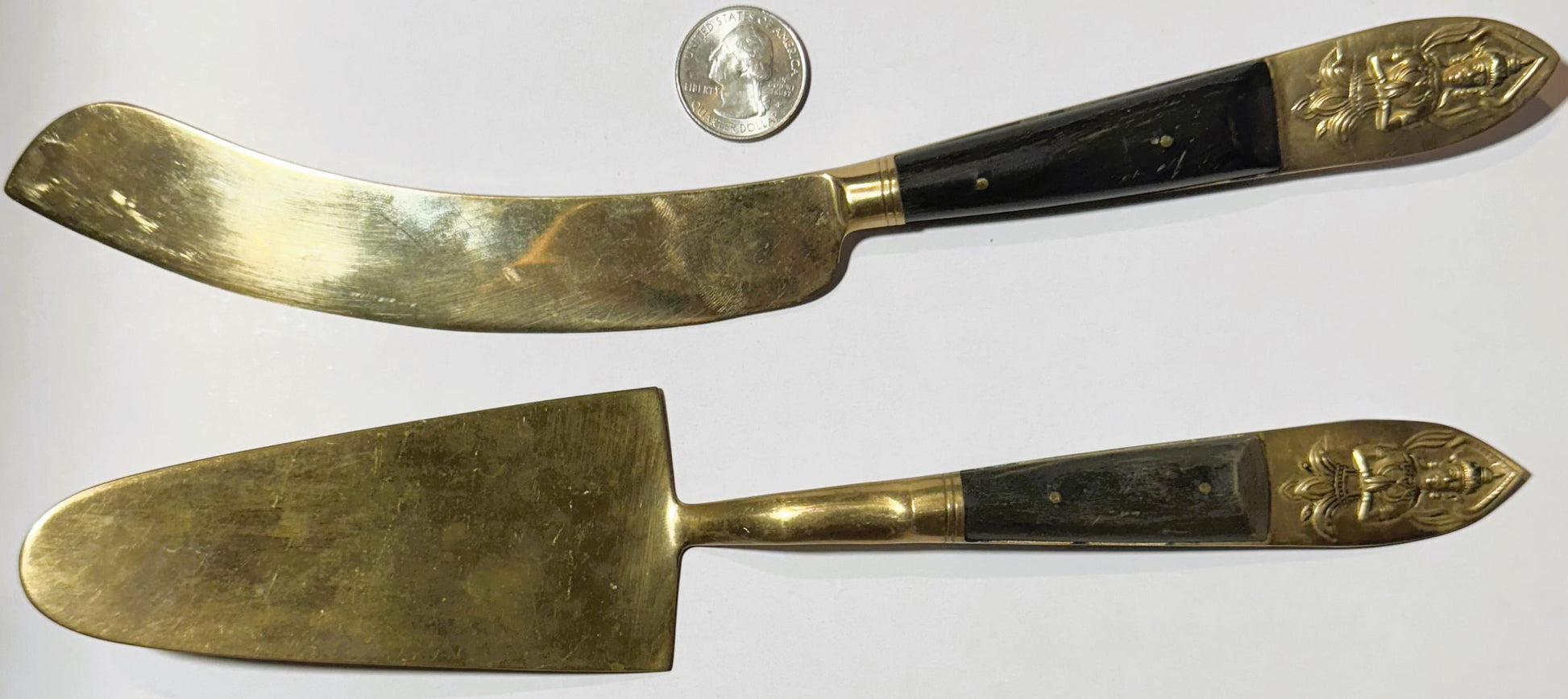 Two brass serving utensils with black handles and ornate deity finials displayed with a quarter for scale on white background