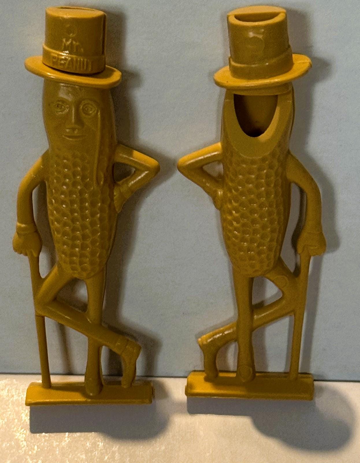 Pair of gold plastic Mr. Peanut figural whistles with top hats, shown front and back views against white background