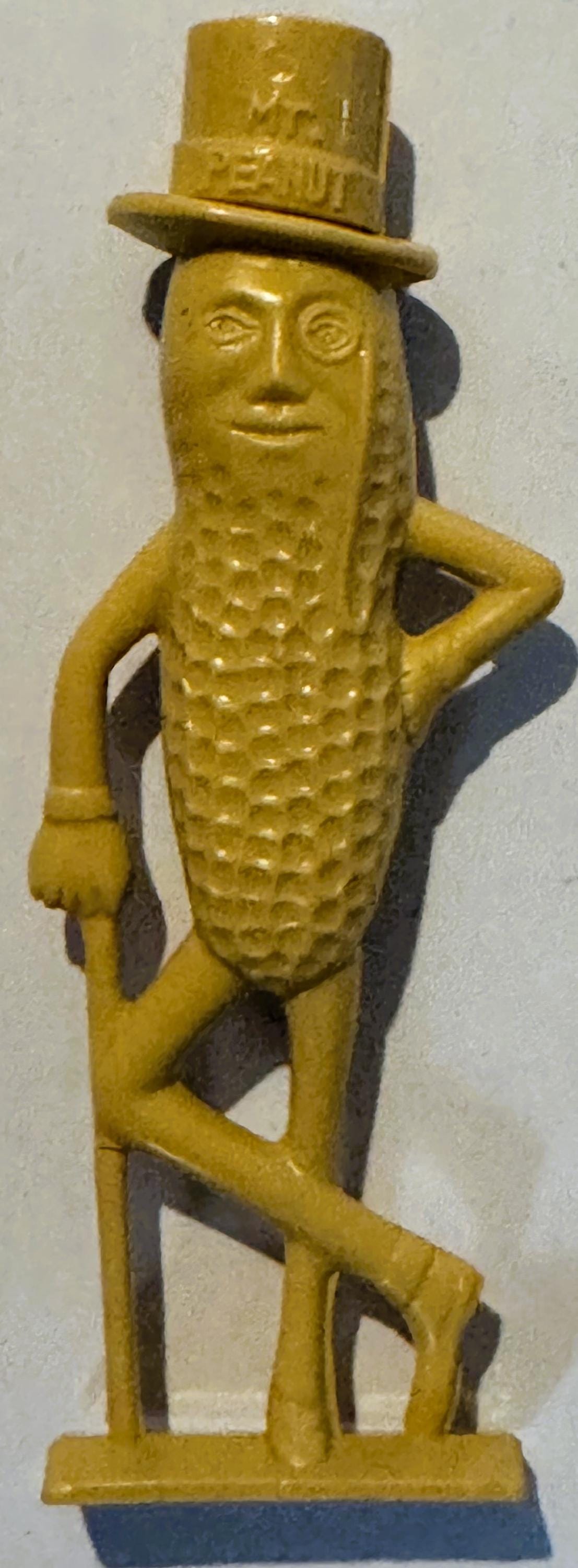 Vintage yellow plastic Mr. Peanut figural whistle with top hat, monocle, and cane, shown standing against white wall with sha