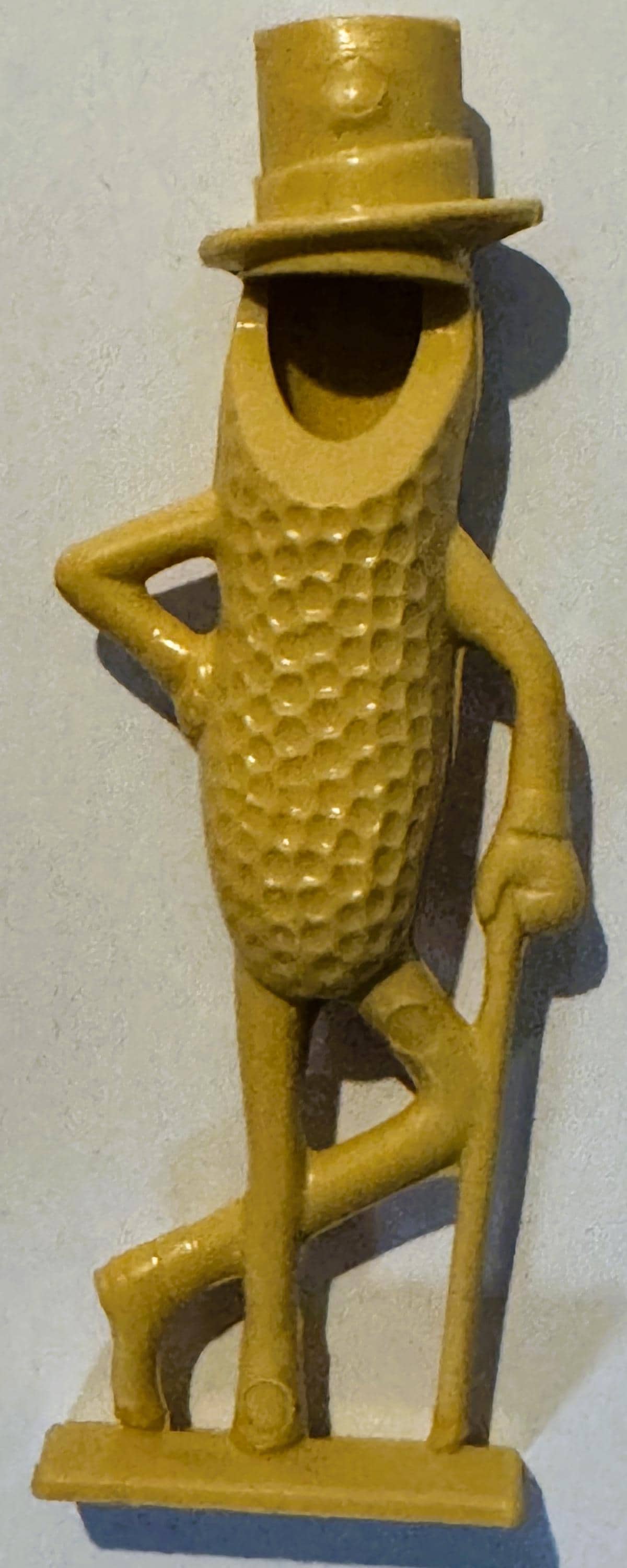 Vintage yellow plastic Mr. Peanut figural whistle with top hat and monocle, standing upright against white wall with shadow v