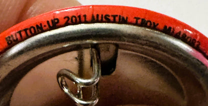 Close-up of red and silver pin held in hand, showing red rim with black text reading "BUTTON UP 2011 AUSTIN TX" and metal