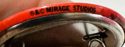 Close-up of red enamel pin rim stamped "C&C MIRAGE STUDIOS" held in someone's hand for scale