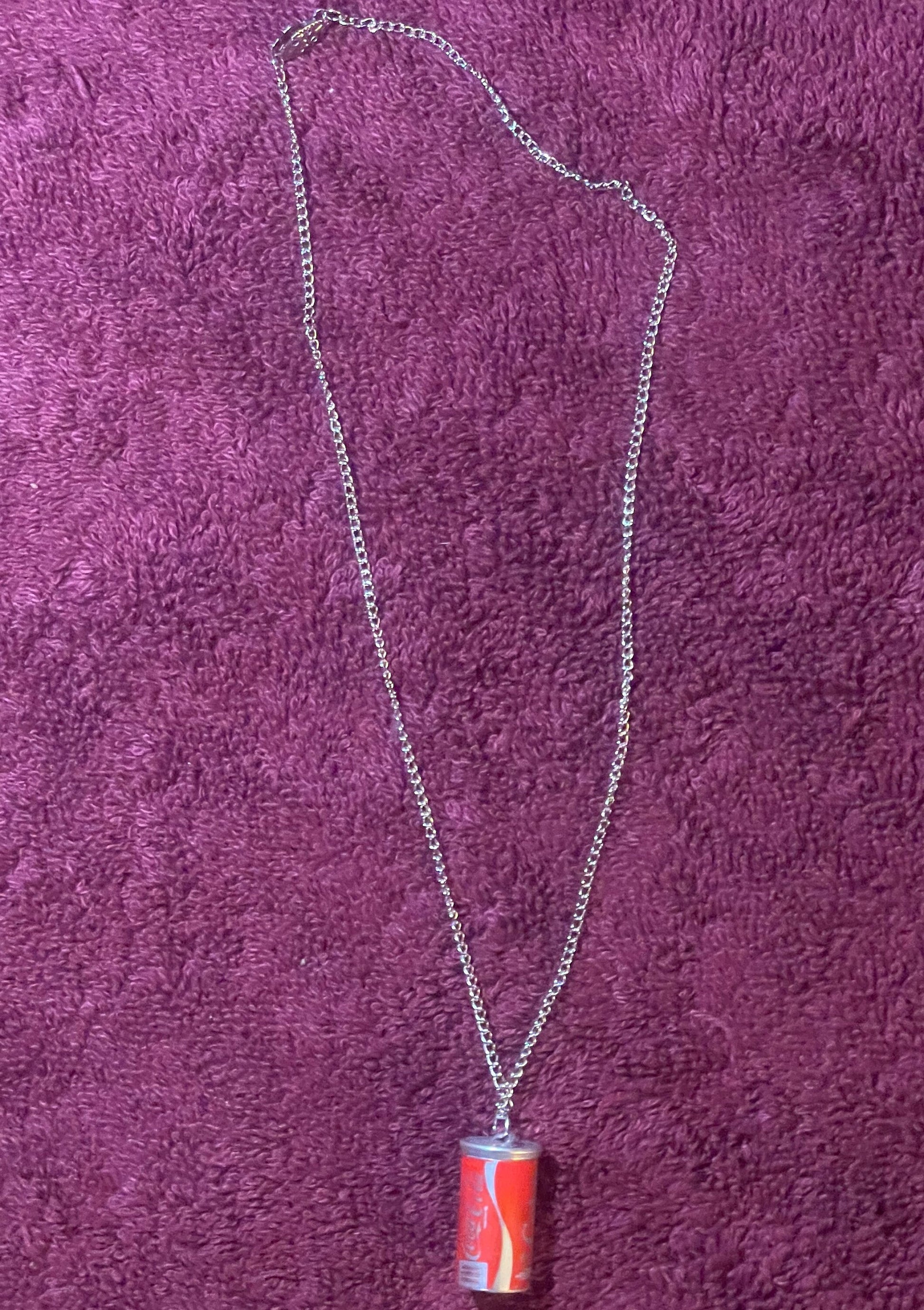 Silver chain necklace with small red Coca-Cola mini can pendant displayed on purple textured fabric background
