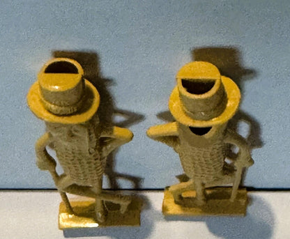 Two yellow and tan plastic Mr. Peanut figurines with top hats facing each other against a light blue wall and white surface