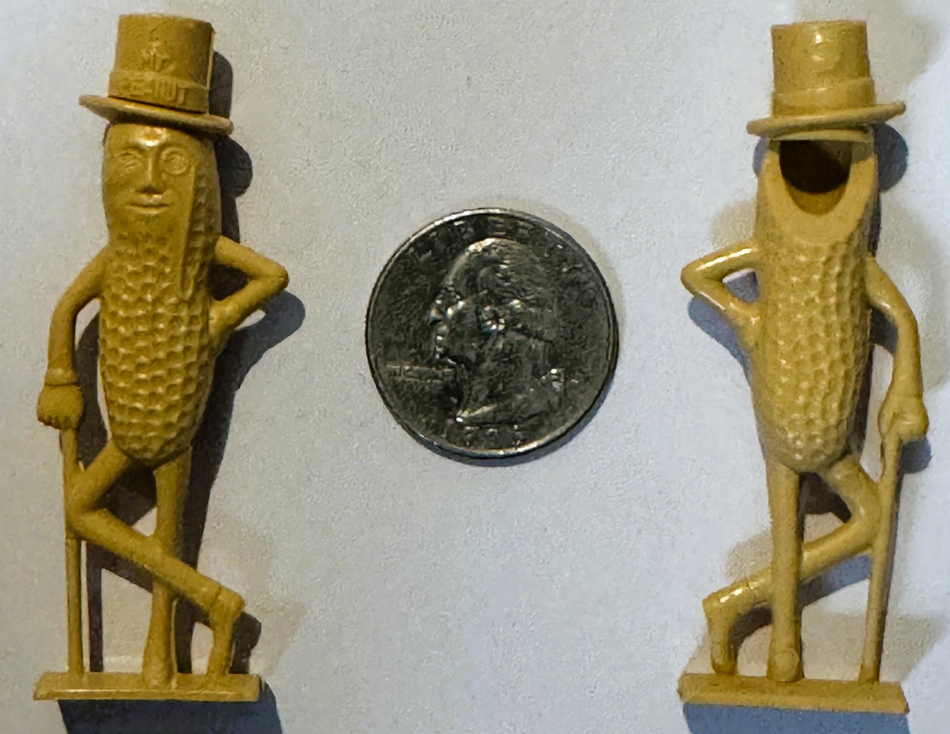 Two yellow plastic Mr. Peanut figurines with top hats flanking a coin for scale against white background