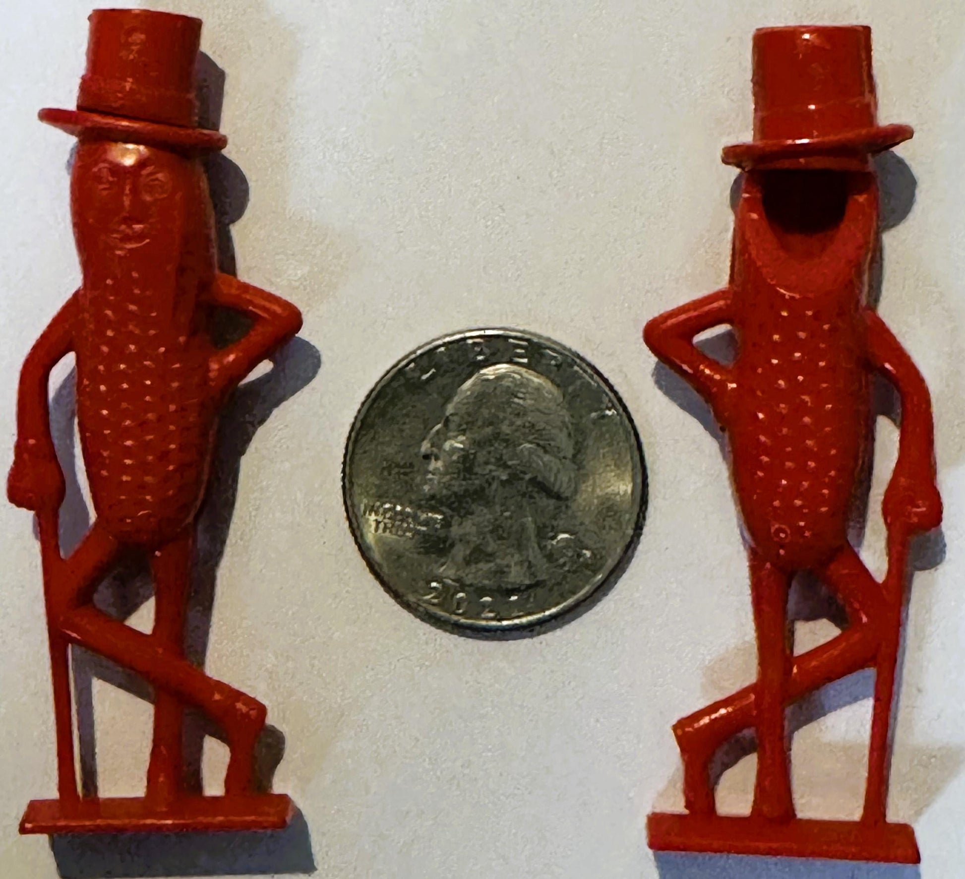 Two red plastic Mr. Peanut figurines with top hats flanking a quarter coin for scale against white background