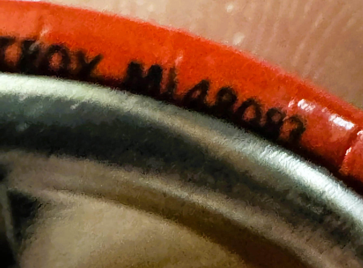 Close-up of red enamel pin with embossed black details on tan fabric, showing partial text and decorative edge design.