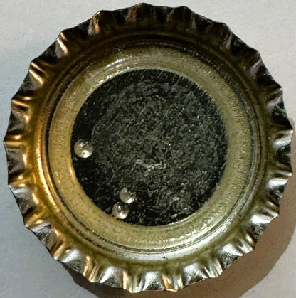 Overhead close-up of a ridged metal bottle cap with brass-colored outer rim and dark center disk with small reflective spots.