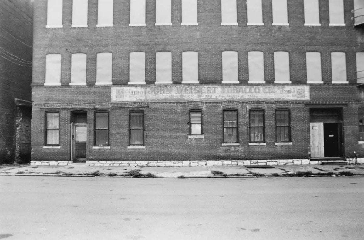 Black and white photograph of a brick building facade with "John Weisert Tobacco Co. St. Louis MO" signage visible above ground-floor windows and doors.