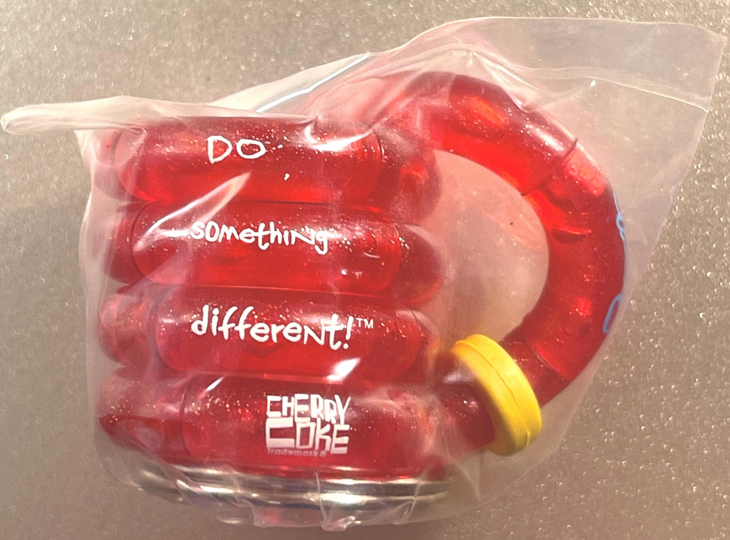 Sealed clear plastic package containing red Cherry Coke branded fidget toy with yellow band, printed text visible including "