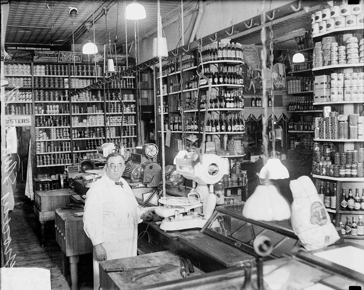 Old black and white photo of a grocery store counter and packed shelves, a grocer in a white coat at the scale - a period photo for atmosphere, not included