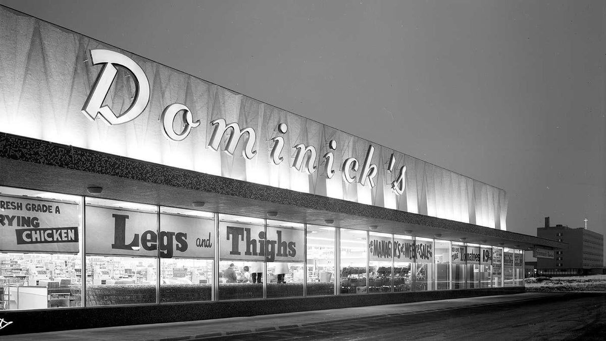 Black and white night photo of a Dominick's supermarket storefront with its lit sign and window signs - a Chicago grocery photo for atmosphere, not included