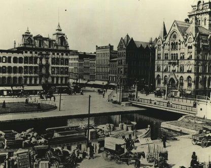 Black and white vintage photograph showing a historic waterfront cityscape with multi-story buildings, a canal, boats, horse-