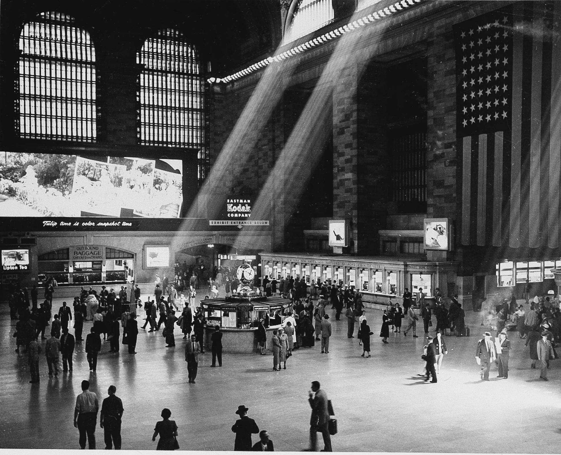 Black and white photograph of Grand Central Terminal's main concourse with crowds of people, dramatic light rays, and America