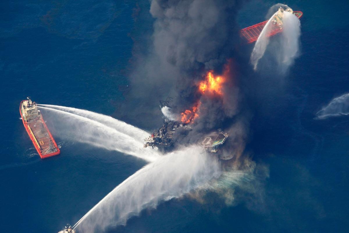 Aerial view of burning offshore oil platform with multiple ships spraying water on intense flames and black smoke