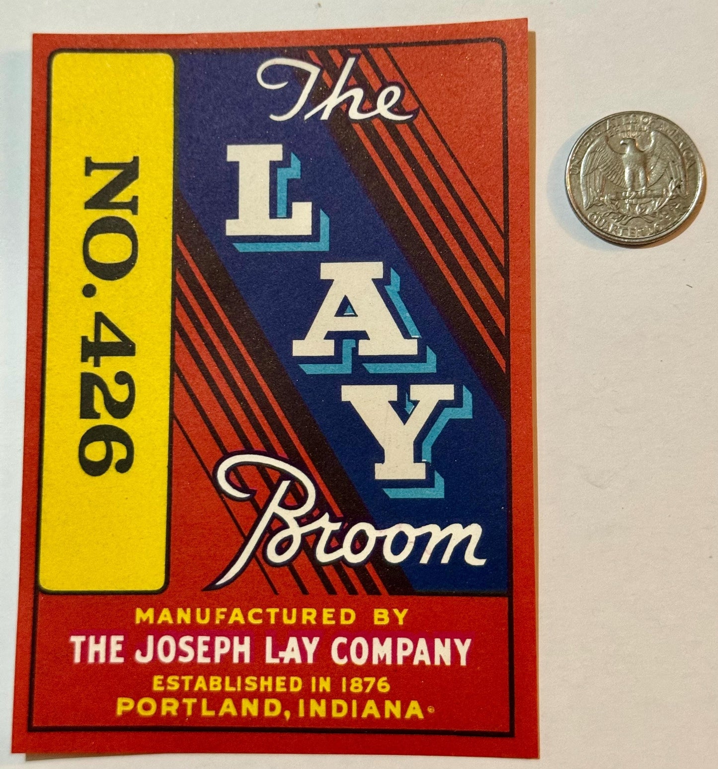 Vintage red, blue, yellow "The Lay Broom" No. 426 label beside a quarter.