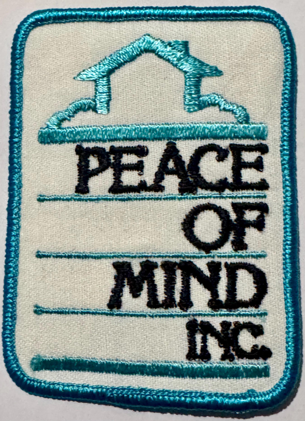 Peace of Mind Inc. embroidered patch: turquoise house outline over a cloud above black PEACE OF MIND INC. lettering on white twill, turquoise merrowed edge