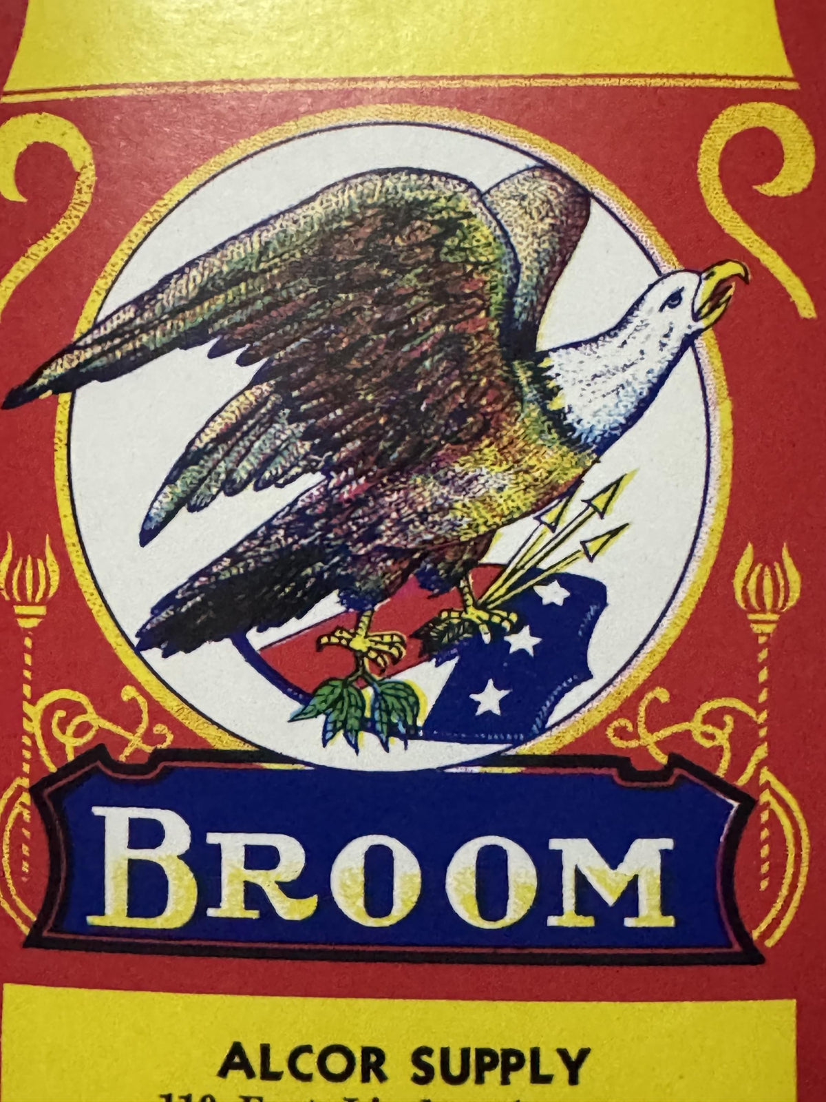 Close-up of the eagle, beak open, gripping arrows and olive leaves on a starred red, white and blue flag, above the BROOM banner
