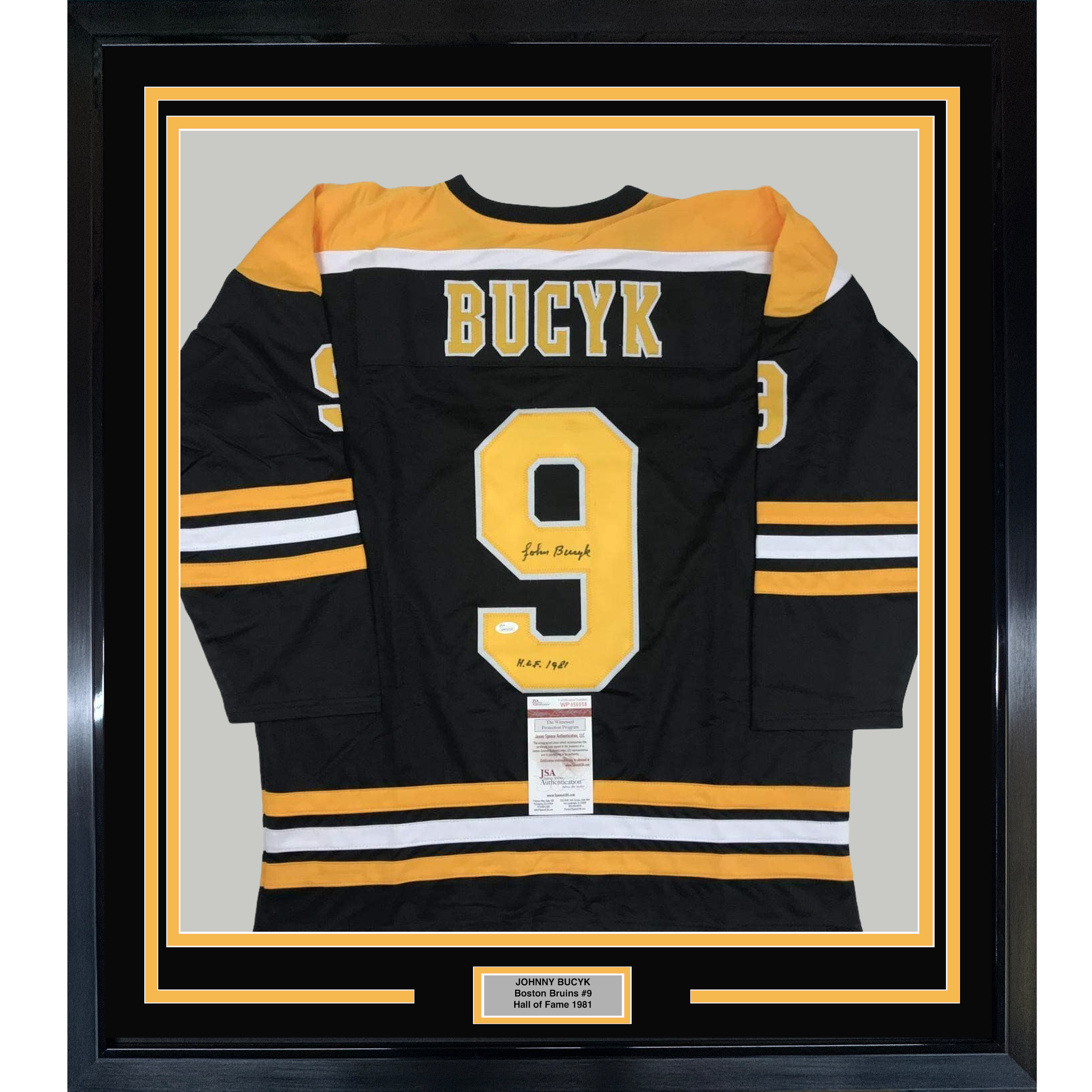 Framed black Boston Bruins hockey jersey number 9 with yellow and white stripes, signed by Johnny Bucyk, displayed flat against gray backing with nameplate below.