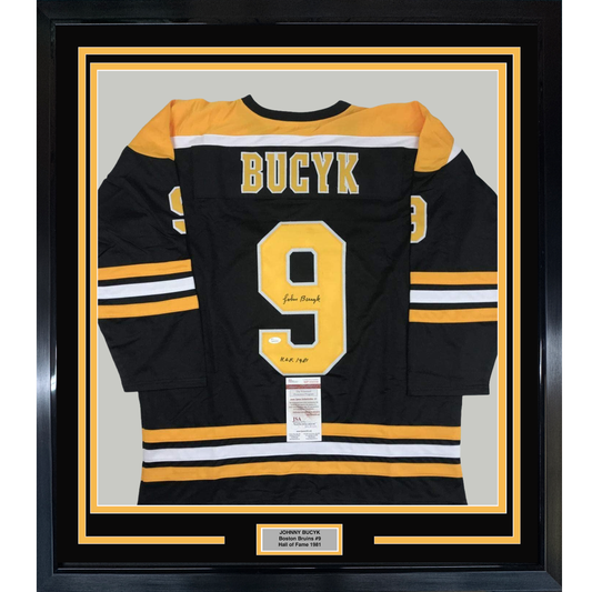 Framed black Boston Bruins hockey jersey number 9 with yellow and white stripes, signed by Johnny Bucyk, displayed flat against gray backing with nameplate below.