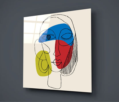 Modern glass wall art displaying an abstract line-drawn face with blue, red, and yellow colored sections mounted at an angle against a gray wall.