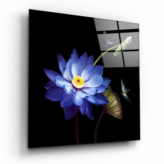 Angled three-quarter view of glass wall art panel displaying blue lotus flower with yellow center and green stems against black background, with dragonflies and small preview grid visible in upper right corner.