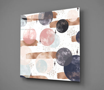 Glass wall art panel with abstract celestial design featuring planets, moons, watercolor brushstrokes in pink, brown, and navy on white background, mounted at angle against gray wall.