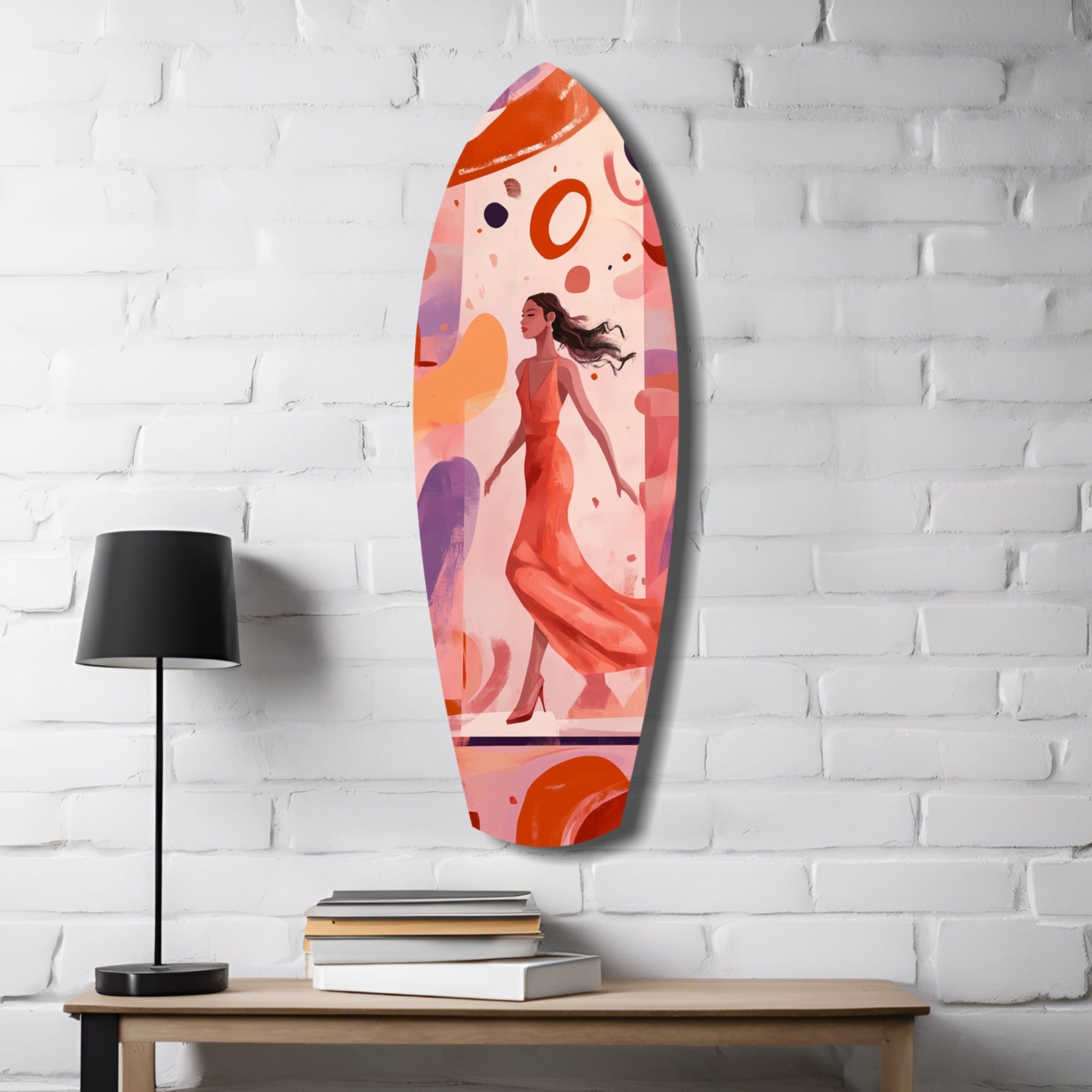 Oval glass wall art featuring illustrated woman in red dress with abstract orange and purple circles on pink background, displayed on white brick wall above wooden table with lamp and books.