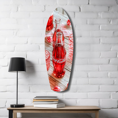 Oval glass wall art featuring a red Coca-Cola bottle on wooden surface with red decorative elements, displayed above wooden desk with lamp and books.