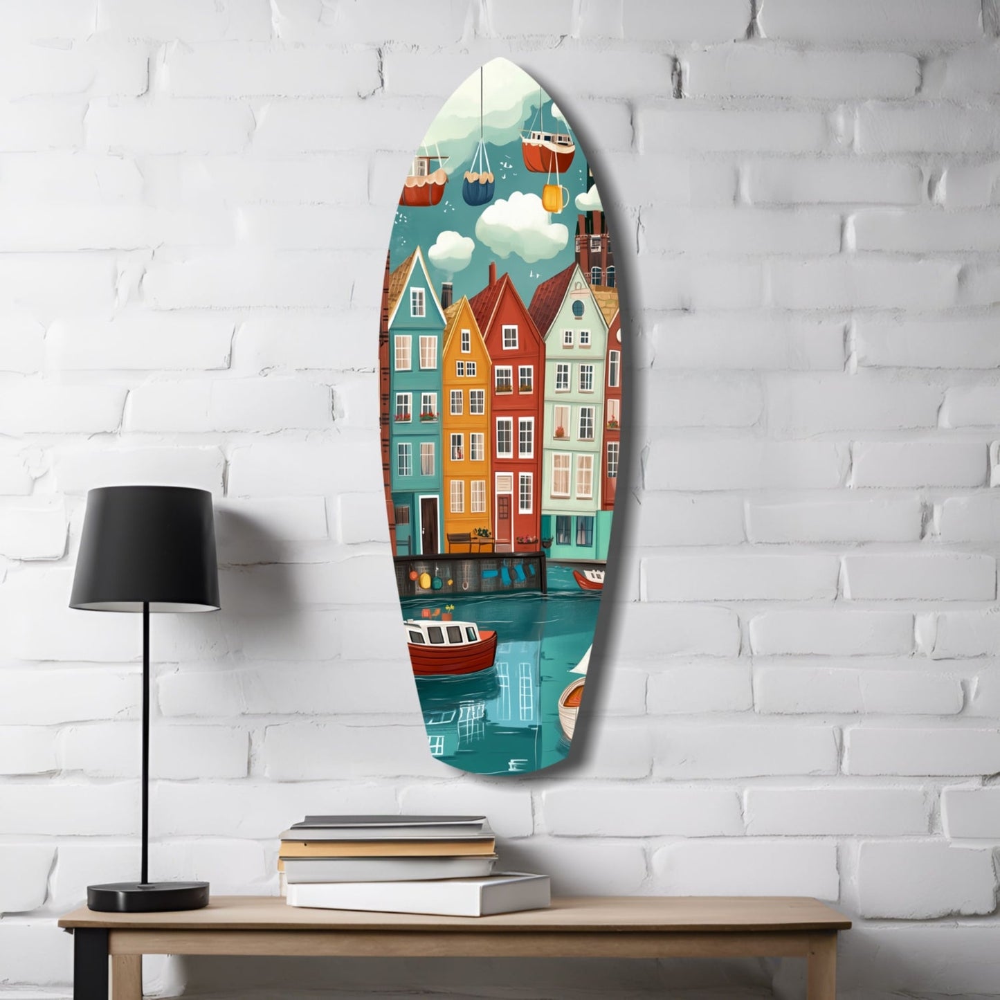 Oval glass wall art featuring colorful coastal townhouses, boats, and sky hanging above wooden table with lamp and books against white brick wall.